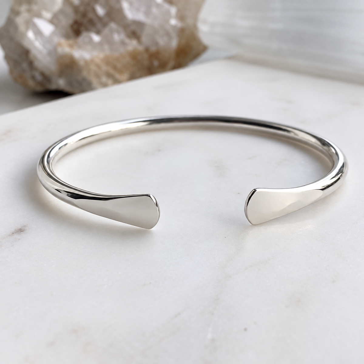 A polished silver open cuff bracelet with a minimalist, tapered end design, resting on a white marble surface with a blurred crystal in the background.