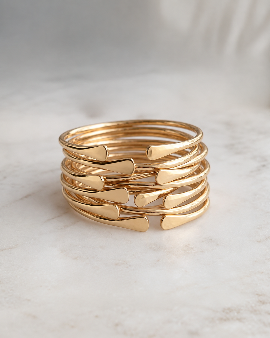 A stack of five polished brass open bangle with flat, curved arrow-like accents, displayed on a light marble surface.