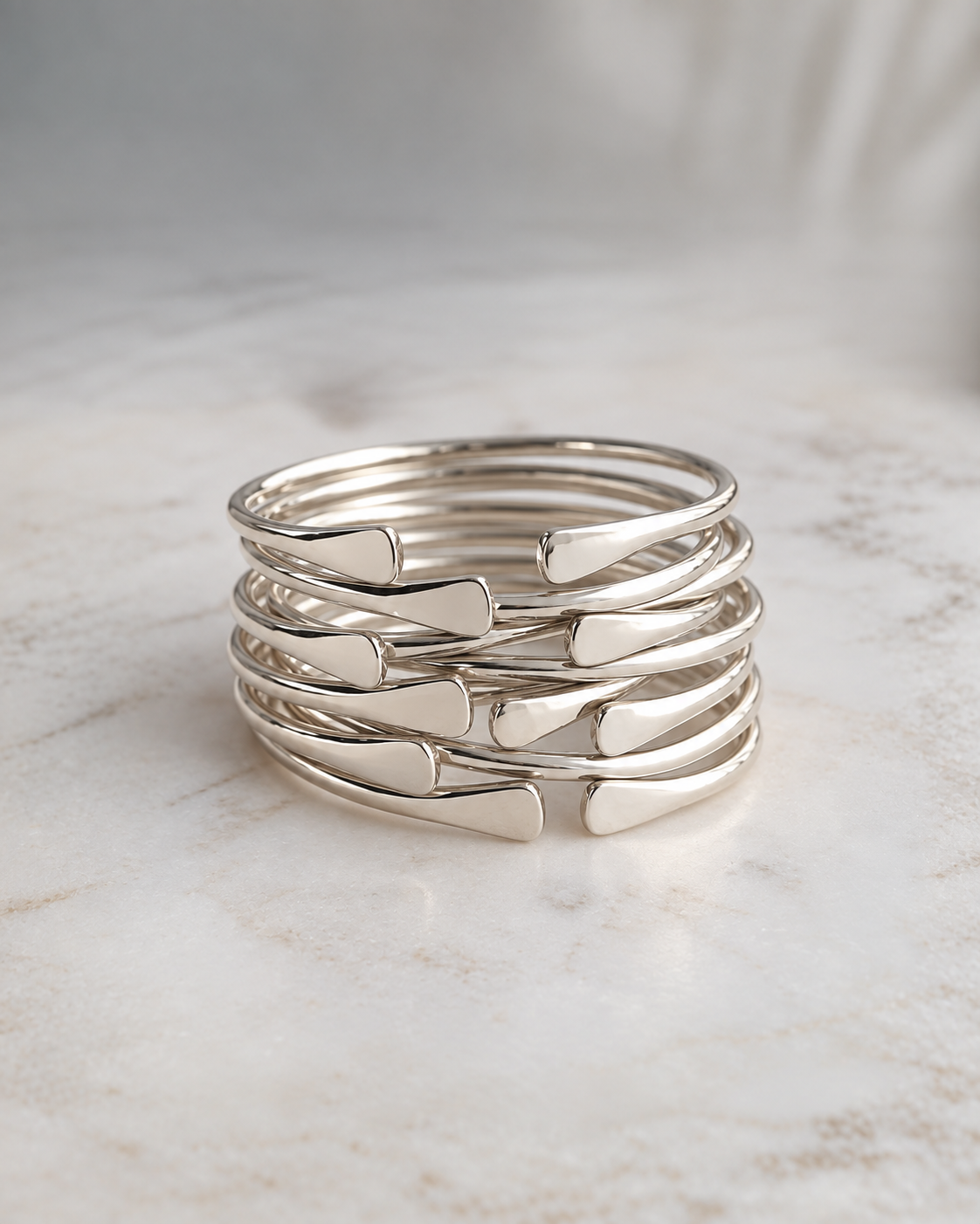 A stack of five polished silver brass bangle bracelets with a twisted, overlapping design, displayed on a light marble surface.