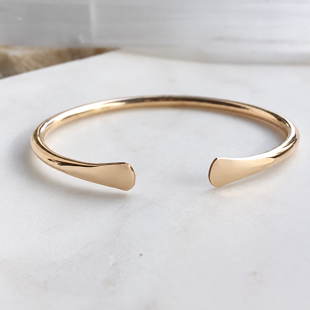 A polished gold-tone brass hand hammered open cuff with a minimalist, tapered design, resting on a light marble surface.